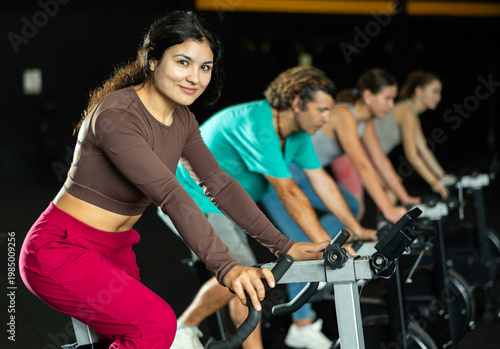 Positive Latina girl training with spin bike in fitness hall during full-body workout