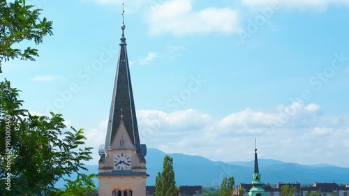 Church clock tower and cityscape against mountains. Beautiful church steeple with a clock tower standing against a clear blue sky with fluffy white clouds, overlooking a picturesque city