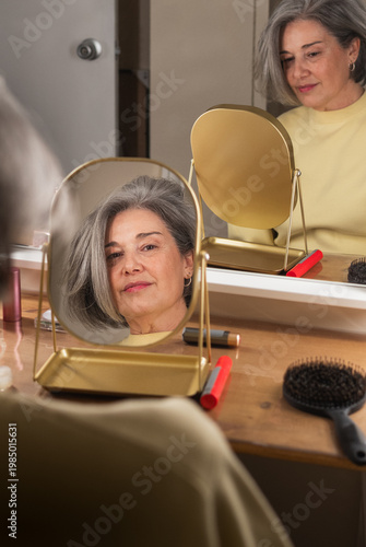 Mature woman looking at her reflection in a gold tabletop mirror, combing gray hair and applying makeup as part of her self-care routine and daily beauty preparation at a wooden vanity