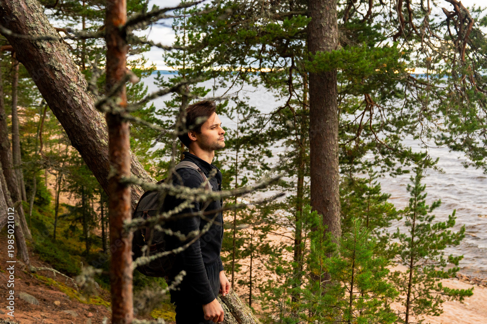 Fototapeta premium Epic northern forest traveler man adventure landscape portrait. Cinematic full length view of male explorer with backpack standing among dense pine trees near wild lake shore water. 