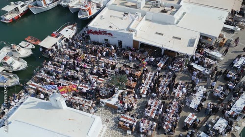 Aerial drone view of Easter celebration in Naousa village on Paros island, Greece, showing people gathered at outdoor tables in a festive Mediterranean atmosphere. Ideal for culture, travel, holidays