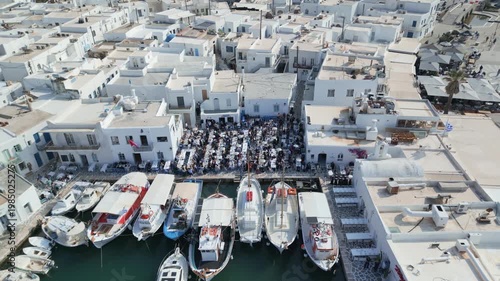 Aerial drone view of Easter celebration in Naousa village on Paros island, Greece, showing people gathered at outdoor tables in a festive Mediterranean atmosphere. Ideal for culture, travel, holidays