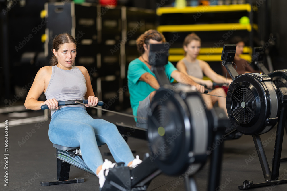 Fototapeta premium Young woman with sports leggings and a top does a cardio workout on a rowing machine. Crossfit group training participants perform a circuit training session for endurance.