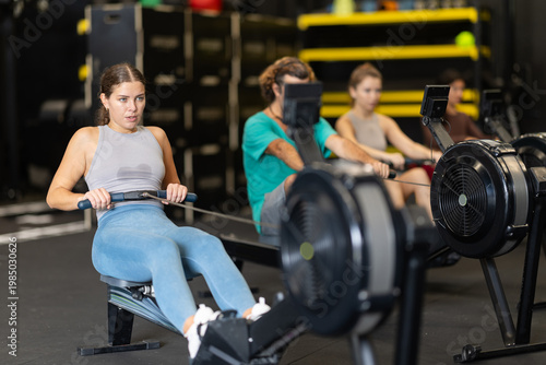 Young woman with sports leggings and a top does a cardio workout on a rowing machine. Crossfit group training participants perform a circuit training session for endurance.
