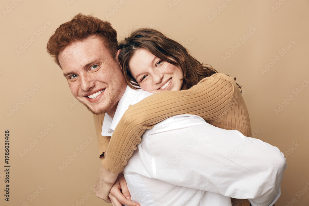 Fototapeta premium Smiling young couple in casual clothes embraces happily against beige background. The woman hugs the man from behind, both showing joyful expressions. Concept of love, friendship, and togetherness.