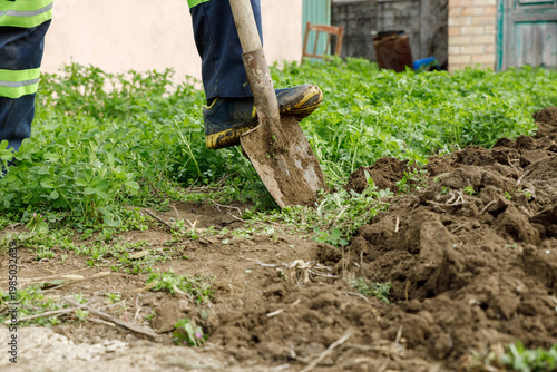 A person is digging soil in a field wearing blue pants and work shoes. The surroundings have green plants.