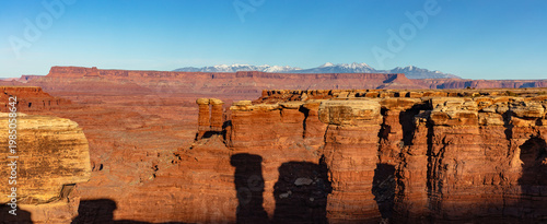 This scenic panorama of Canyonlands National Park features sandstone pinnacles along the rim of Gooseberry Canyon with the snow-capped La Sal Mountains in the distance under a blue sky.