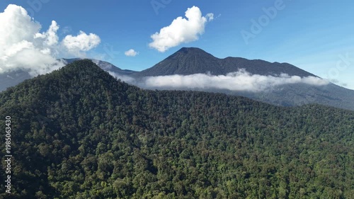Aerial view of dense tropical rainforest covering mountain slopes with low clouds drifting across the peak under a bright blue sky in Indonesia.