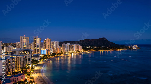 Vibrant Honolulu city skyline and Waikiki Beach illuminated at twilight, with Diamond Head rising majestically against the deep blue sky.
