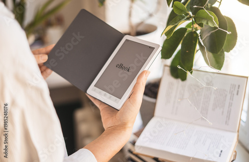 Close view of female hands using ebook device at home. Warm sunlight, minimal interior and green plant create calm reading environment, combining technology and comfort in everyday lifestyle