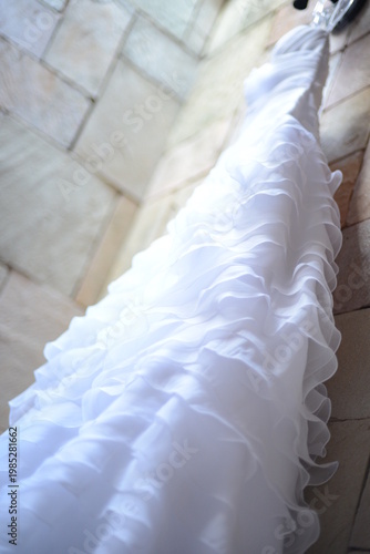 white wedding dress, elegant white ruffled wedding dress hanging against stone wall, vertical view of bridal gown with layered organza ruffles, beautiful wedding dress preparation on stone background