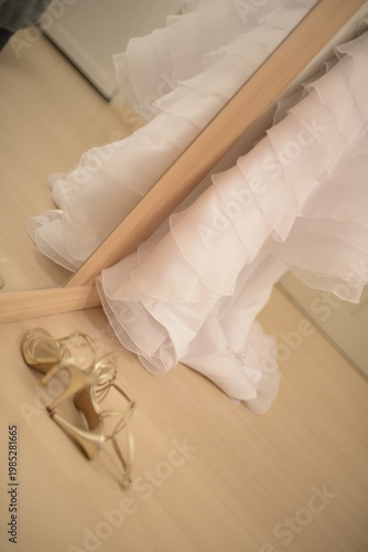 golden bridal sandals next to a white ruffled wedding dress reflection in the mirror, elegant wedding shoes and gown preparation detail, gold high heels and layered bridal dress in room setting, weddi