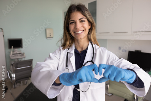 Foto Portrait of happy Latina cardiologist woman in white coat wear blue rubber protective gloves showing symbol of love, promoting regular heart check up for cardiovascular disease prevention