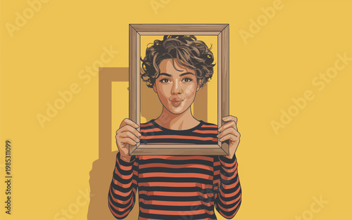 A young man holding a picture frame in front of his face smiles playfully indoors.
