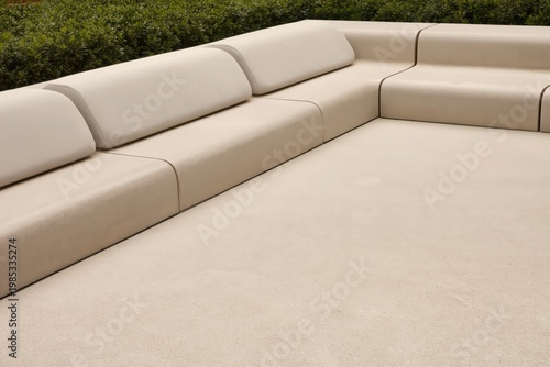 Sectional sofa featuring a modular design with beige upholstery is positioned against a low wall in a modern outdoor lounge area surrounded by a neatly trimmed green hedge.