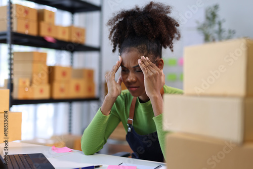 Schilderij op canvas Stressed African American entrepreneur holding her head in pain, surrounded by cardboard boxes in a chaotic office, embodying burnout and exhaustion