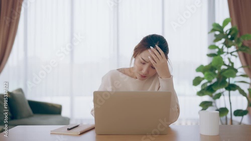 Young Asian woman working in living room, she has a headache due to long and overworking leaving her tired and dizzy. Concept of sickness from work and office syndrome