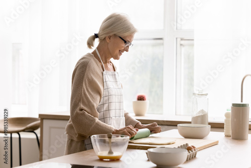 Beautiful European aged woman wear apron flattening dough, cooking, making homemade cake for family on holiday in kitchen. Food preparation, household chores, untroubled retirement and culinary hobby