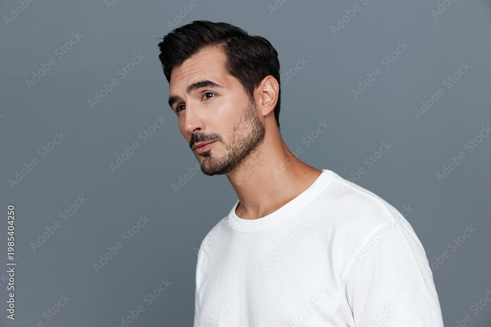 Fototapeta premium Handsome man wearing a white t-shirt looking thoughtfully to the side against a soft grey background, showcasing a contemplative and stylish vibe.
