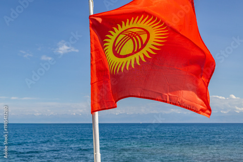 Flag of Kyrgyzstan proudly flutters on pole against a backdrop of calm Issyk-Kul lake and bright sky. Fluffy clouds add to the serene atmosphere of the day.