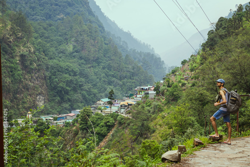 Canvas Print A man with a backpack stands on a mountain path overlooking a small village in t