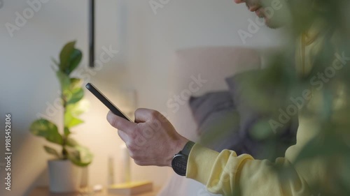 Man using smartphone while sitting in bedroom with houseplant and soft light. Concept of mobile technology, online communication and home lifestyle.