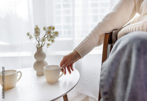 Young woman sitting indoors and drinking coffee or tea in a cozy modern home. Natural light from window, relaxed lifestyle moment with soft atmosphere. Warm tones, minimal interior design.