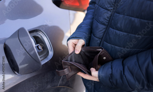 Man sitting in car at gas station during fuel crisis,checking empty wallet and holding payment card.Concept of rising fuel prices.Driver leaning on steering wheel.Close of fuel nozzle into car tank.