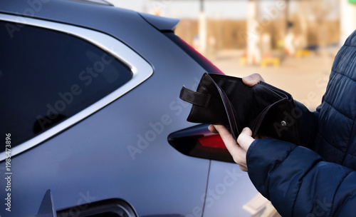 Man sitting in car at gas station during fuel crisis,checking empty wallet and holding payment card.Concept of rising fuel prices.Driver leaning on steering wheel.Close of fuel nozzle into car tank.