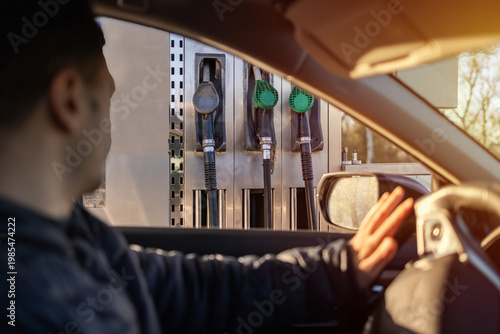 Man sitting in car at gas station during fuel crisis,checking empty wallet and holding payment card.Concept of rising fuel prices.Driver leaning on steering wheel.Close of fuel nozzle into car tank.