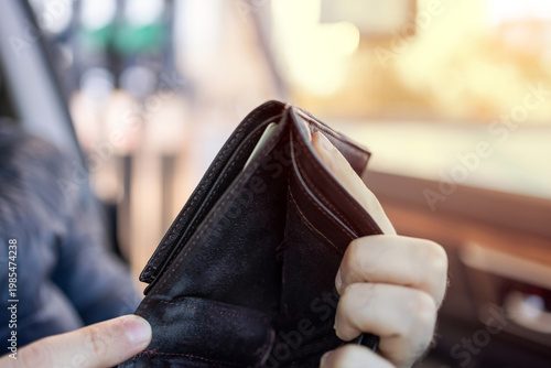 Man sitting in car at gas station during fuel crisis,checking empty wallet and holding payment card.Concept of rising fuel prices.Driver leaning on steering wheel.Close of fuel nozzle into car tank.