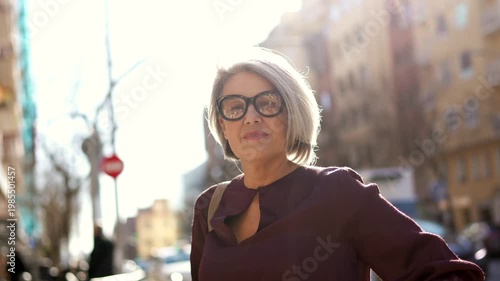 Elegant mature woman with stylish grey hair and glasses standing confidently on a sunny city street, looking at the camera with a slight smile and her arms crossed on a beautiful day