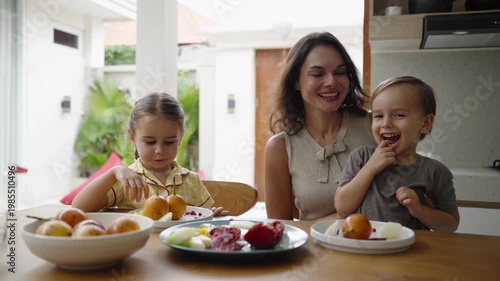 Close up of happy family having breakfast together: mother and two children laughing, hugging, and sharing warm moments as mom gently kisses her kids at the table.