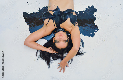 Sexy nude young woman lying on her back with black paint splattered across her chest and the surface, creating a bold artistic and conceptual visual against a white background