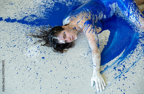 A young woman lies on the floor, her body and hair covered in bold splashes of cobalt blue and white paint. The dynamic scene merges human form with expressive color and movement