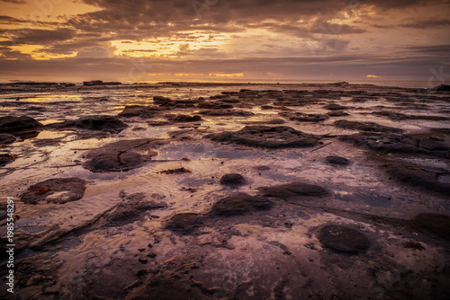 Light reflections from a cloudy sunrise over the sea light up puddles and rocks on an almost flat rock platform formed by sea erosion near the river mouth at Burrill Lake, New South Wales, Australia.