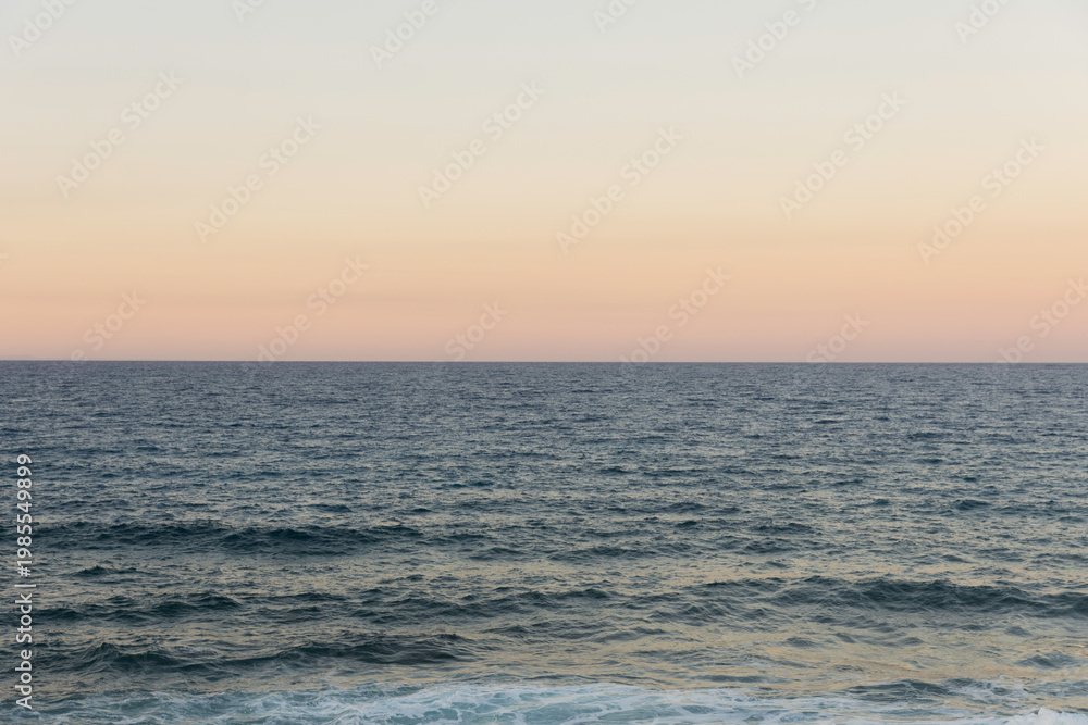 Obraz premium View of the ocean at sunset with gentle waves and a clear horizon under a pastel sky