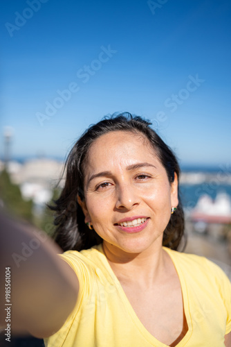 Latin woman smiling at the camera, capturing a selfie during outdoor travel in a sunny destination