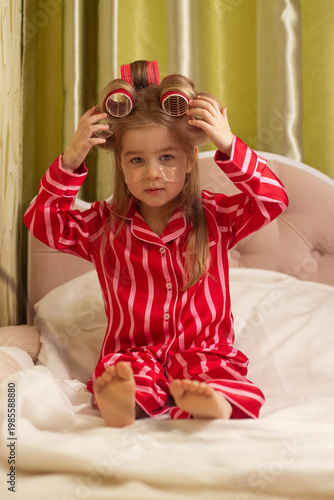 Cute little girl in red striped pajamas sitting on bed with hair rollers and under-eye patches. Morning beauty routine concept, skincare, self-care, childhood lifestyle, cozy home interior