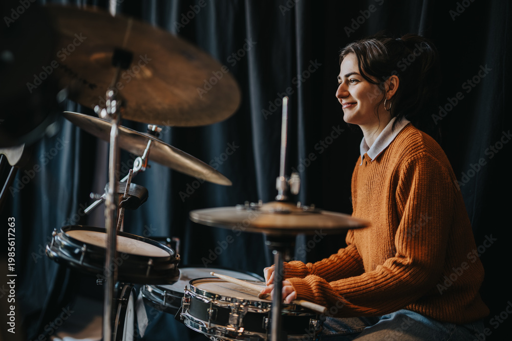 Obraz premium A young woman student plays a drum kit, smiling as she holds drumsticks. She rehearses on snare and cymbals in a dimly lit practice space.
