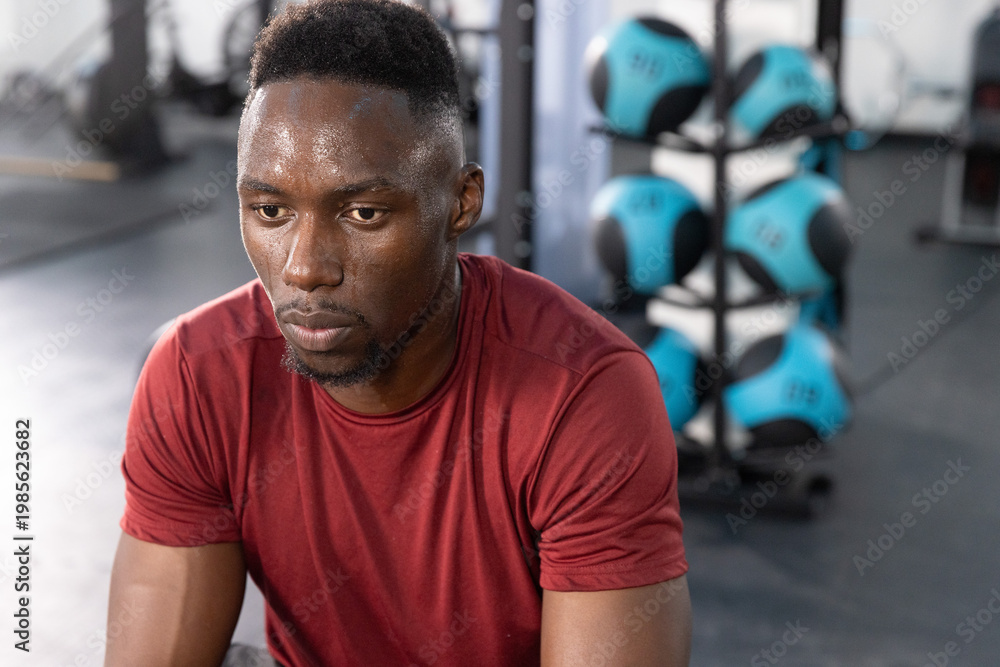 Obraz premium Adult African American man sitting at fitness center in burgundy tee, studying medicine ball rack