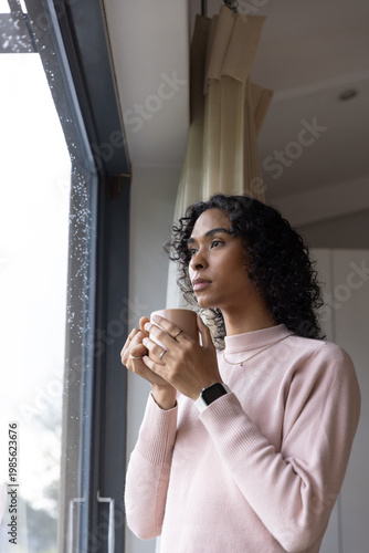 Non-binary adult standing by rainy window holding ceramic mug in pink sweater, showing smartwatch