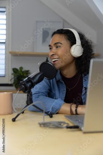 Non-binary podcaster recording at wooden desk in loft studio with microphone, headphones and laptop