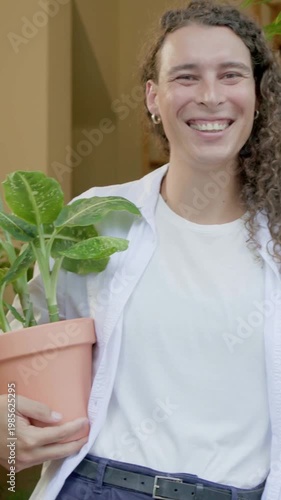 Vertical video: Smiling nonbinary adult holding pot in doorway while camera zooming, sharing plant