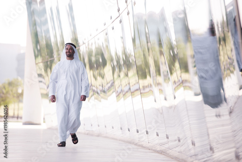 Smiling Arab man in traditional dress walking by reflective modern architecture in an urban park