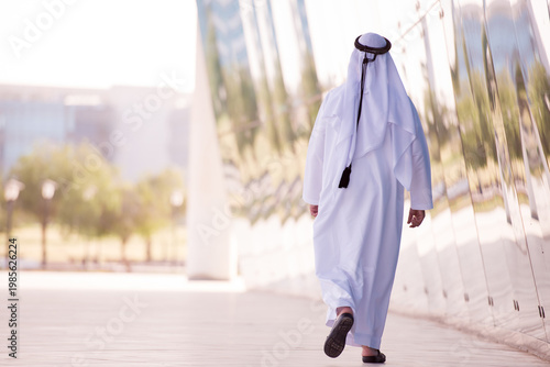 Man in white thobe walking by reflective modern building facade in an urban setting