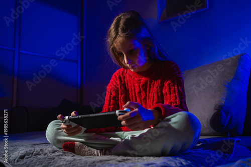 Focused child playing a handheld game console in a dark bedroom lit by blue ambient light