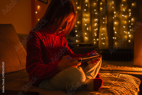 Girl in a red sweater playing a portable game device on a bed illuminated by warm string lights
