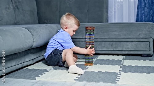A young child sits on a play mat in a living room and concentrates on stacking colorful blocks into a tall tower. The couch is gray and the window is covered with curtains.