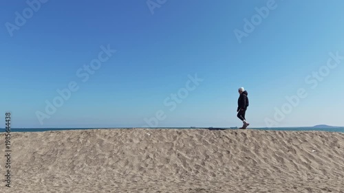 A person walks along a sandy beach with ocean in the background. The sky is clear and the sun is shining. This scene shows a typical day at the coast.